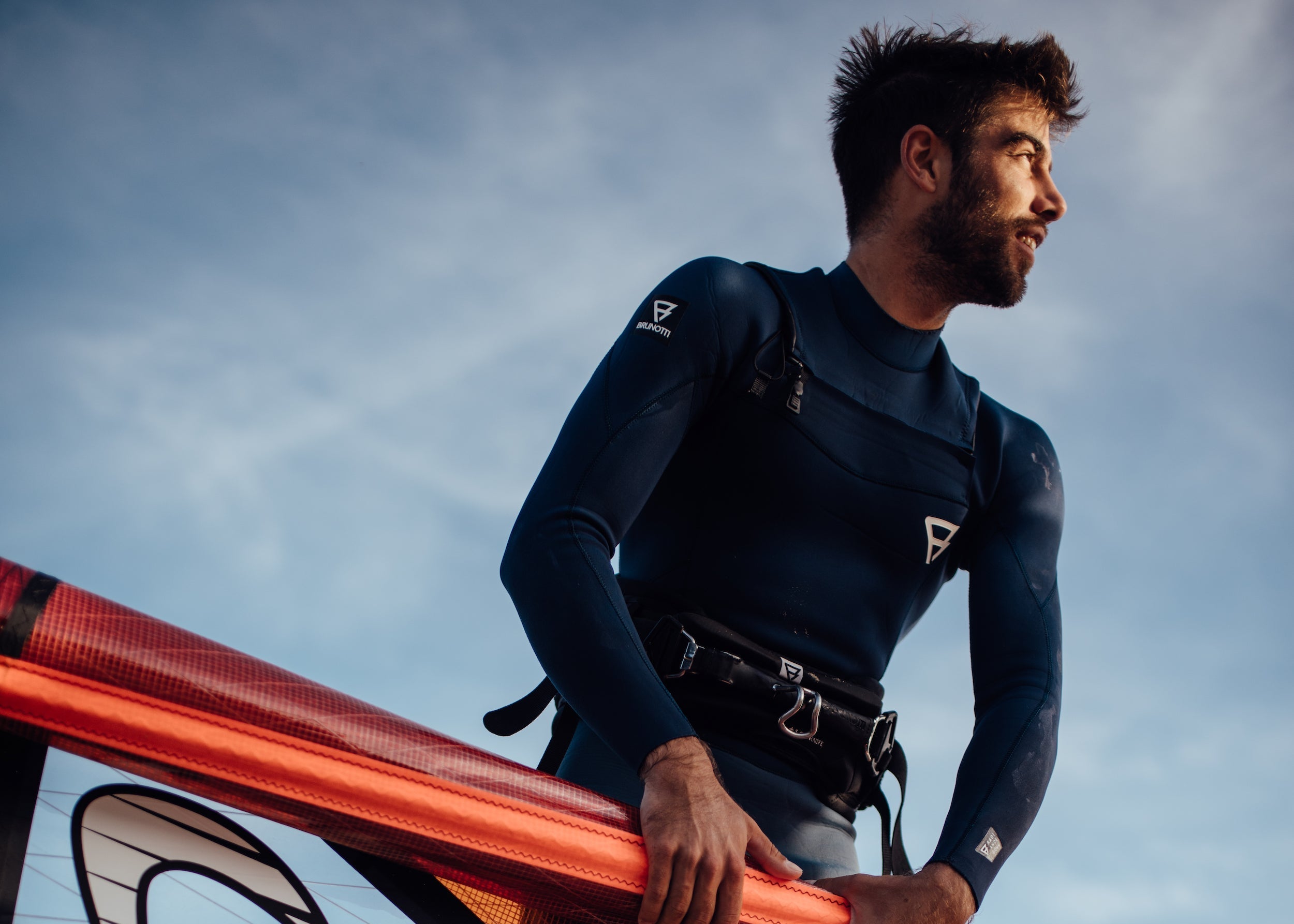 Person in a wetsuit holding an orange windsurf board against a cloudy sky.