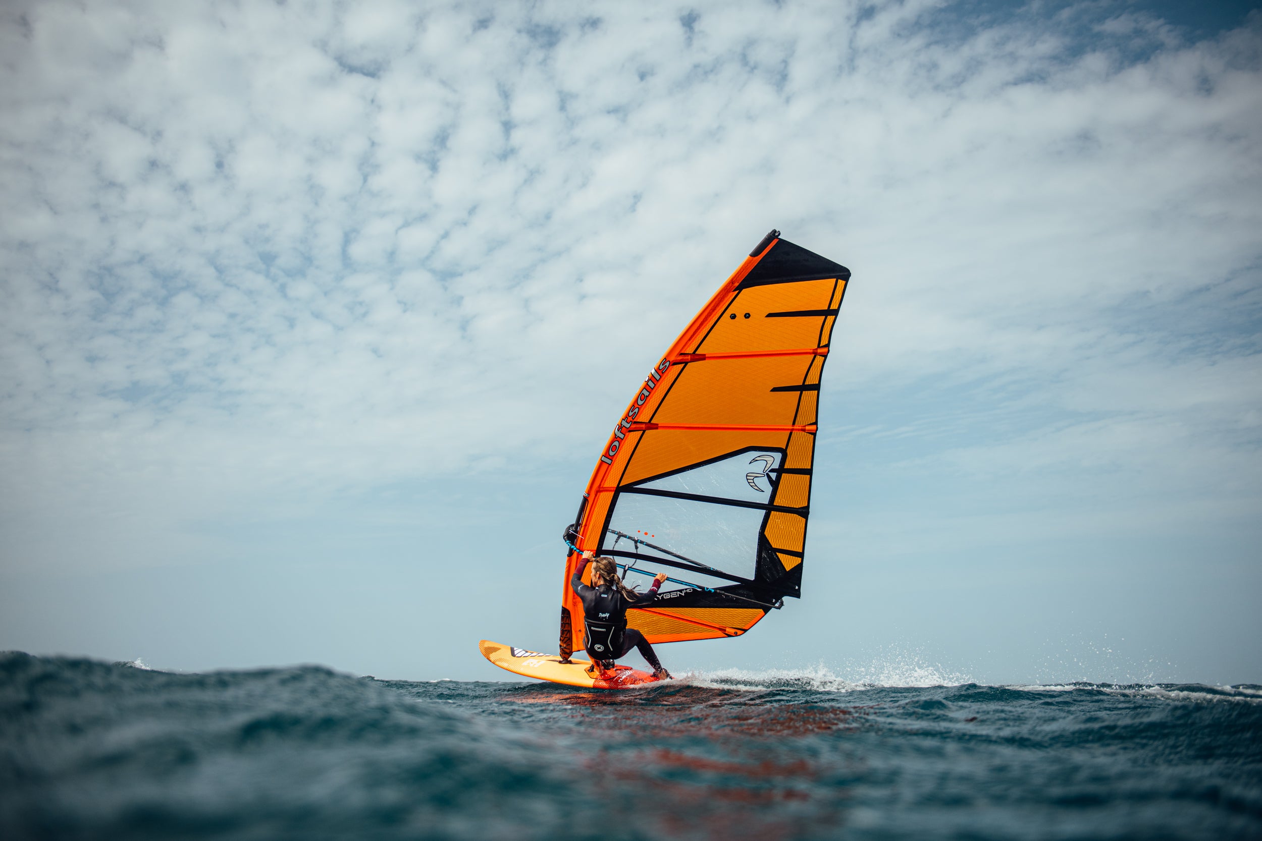 Person windsurfing on a wave with an orange sail against a blue sky.