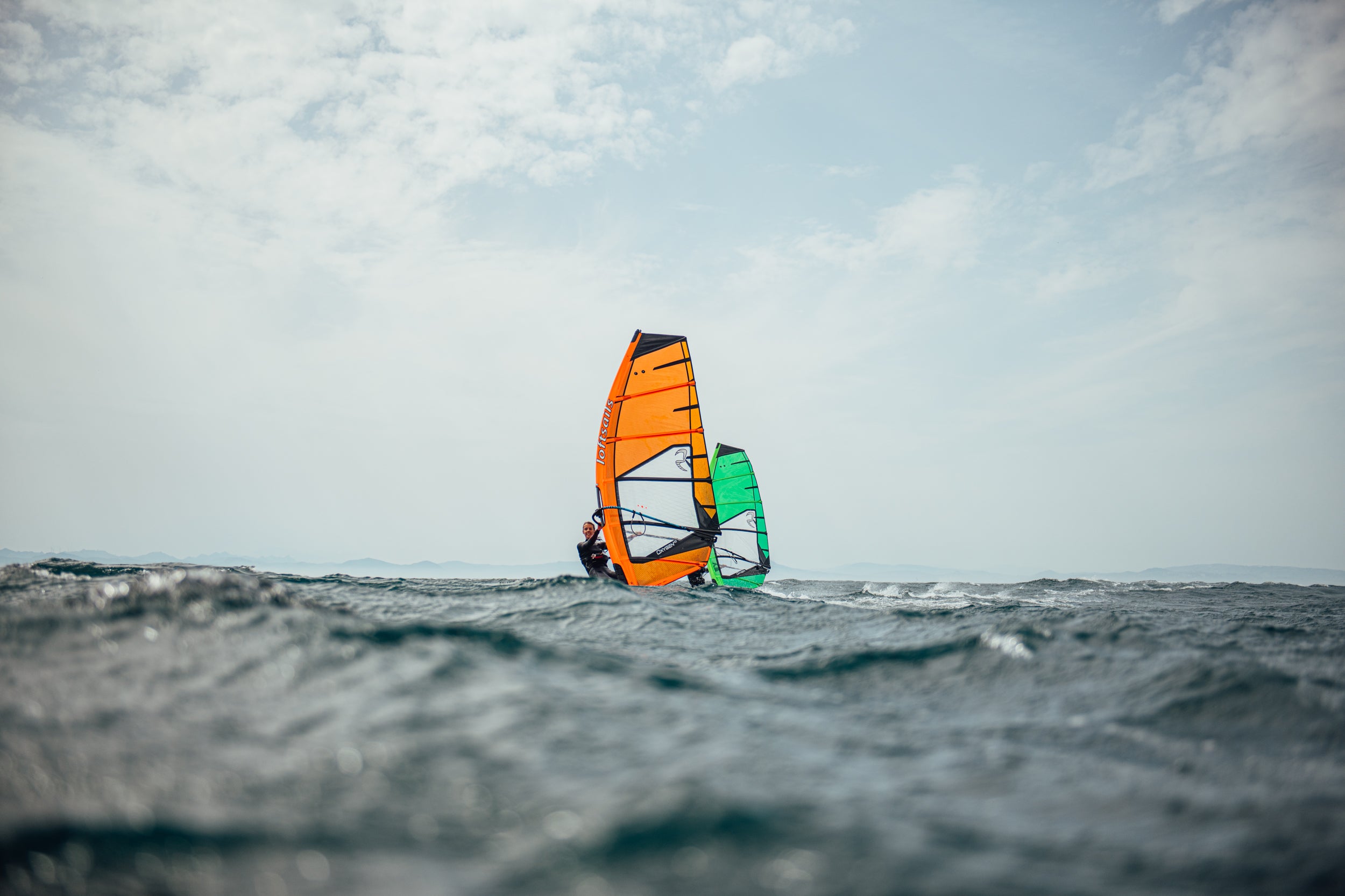 Windsurfer with an orange and green sail on a body of water under a cloudy sky.