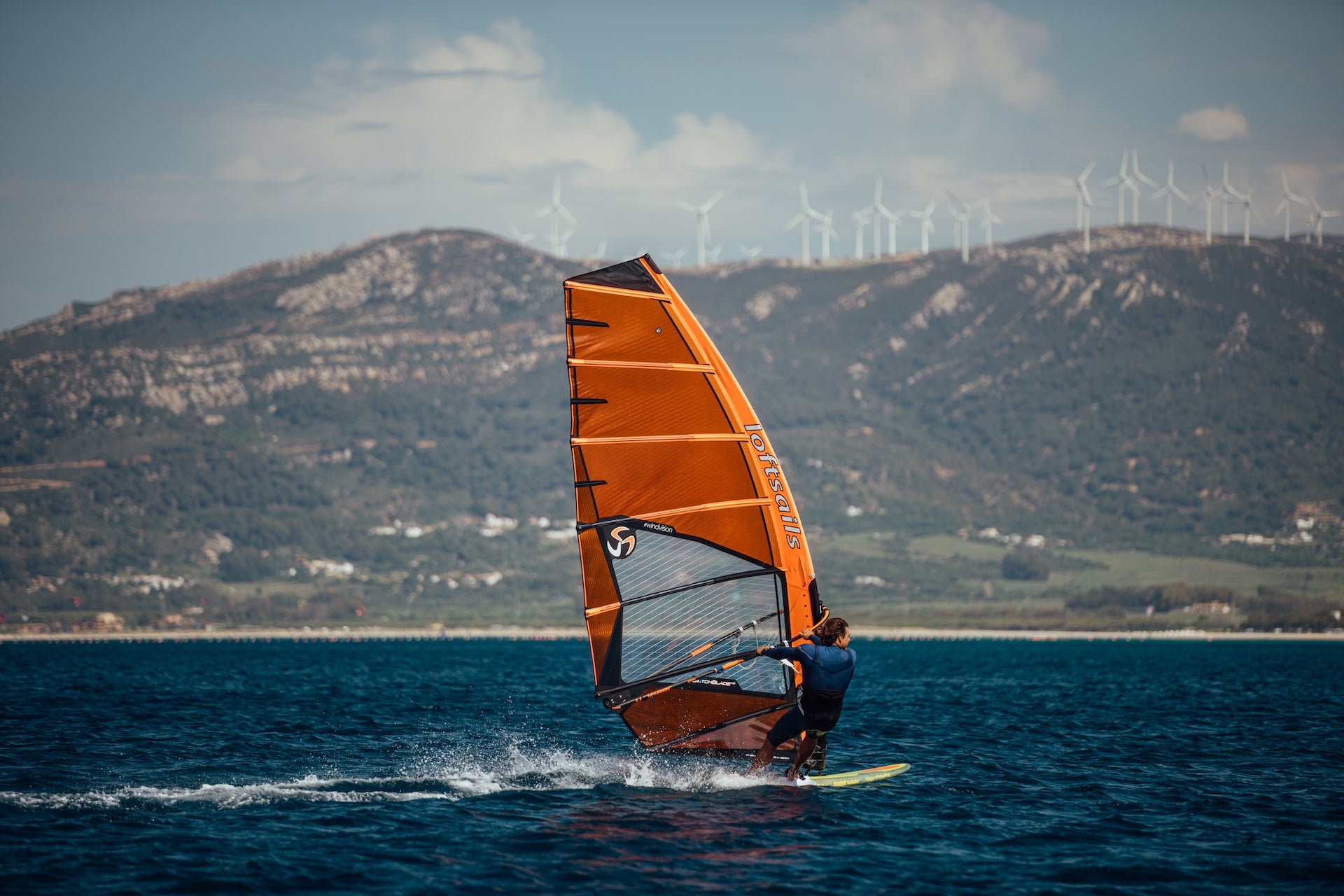 Person windsurfing on a blue sea with mountains and wind turbines in the background