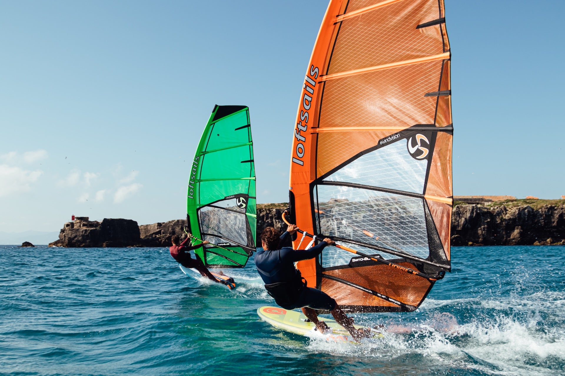 Two windsurfers on the water with colorful sails against a clear sky.